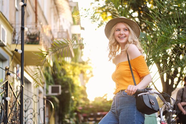 Bastante alegre chica rubia con sombrero felizmente mirando hacia otro lado sobre la puesta de sol en la calle de la ciudad