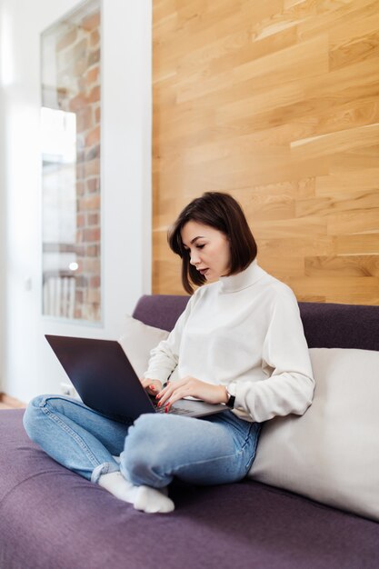 Bastante casual mujer vestida con jeans y camiseta blanca está trabajando en la computadora portátil en casa