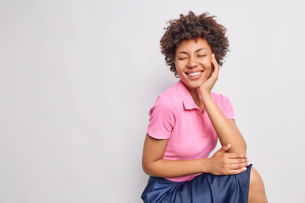 Bastante feliz joven afroamericana de pelo rizado sonríe con dientes mantiene los ojos cerrados vestidos con camiseta rosa casual y falda aislada sobre pared blanca