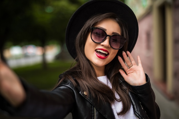 Bastante hermosa mujer asiática sonriente con sombrero y gafas de sol tomando selfie en calle de la ciudad