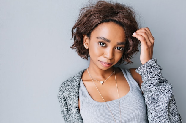 Foto gratuita bastante joven africana con peinado corto y rizado en pared gris. vistiendo chaqueta de punto gris, chaqueta de punto, camisa, collar con triángulo y elegantes pendientes.