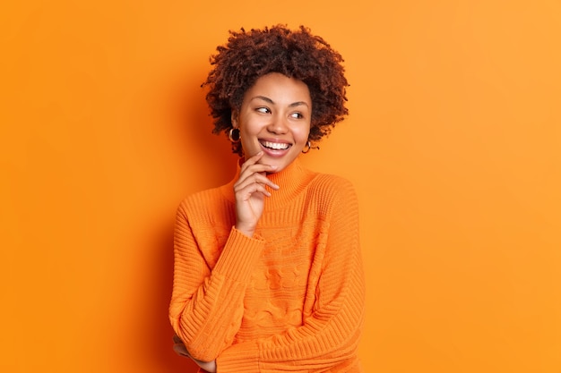 Bastante joven afroamericana mira a un lado con una sonrisa dentuda y nota que algo agradable tiene una expresión despreocupada vestida con un jersey informal posa interior contra una pared naranja vívida