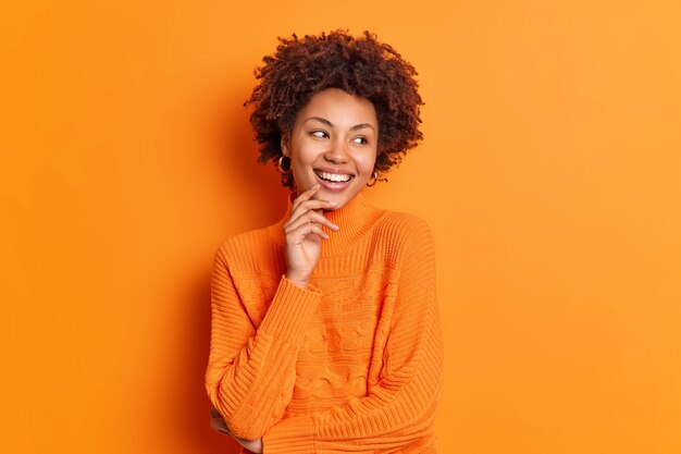 Bastante joven afroamericana mira a un lado con una sonrisa dentuda y nota que algo agradable tiene una expresión despreocupada vestida con un jersey informal posa interior contra una pared naranja vívida