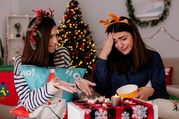 Bastante joven ansiosa con diadema de renos mira palomitas de maíz caídas sentado en un sillón con un amigo tiempo de Navidad en casa