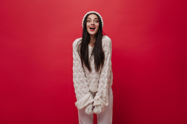 Bastante joven con cabello largo morena labios rojos gorro de punto blanco y guantes pullover sonriendo y posando en traje de Navidad contra el fondo rojo.