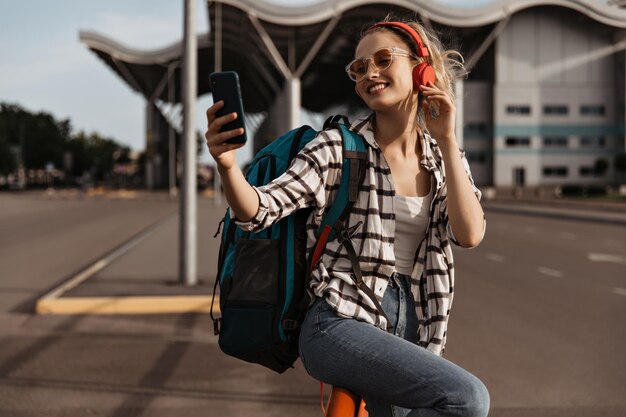 Bastante joven en camisa a cuadros se toma selfie y sonríe Turista sostiene teléfono y mochila Chica rubia con gafas de sol escucha música en los auriculares