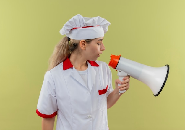 Bastante joven cocinero en uniforme de chef mirando hacia abajo y hablando por altavoz aislado sobre fondo verde