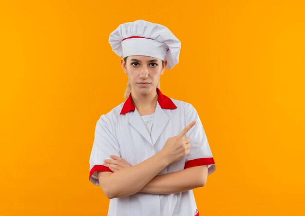 Bastante joven cocinero en uniforme de chef de pie con postura cerrada y apuntando a un lado y mirando