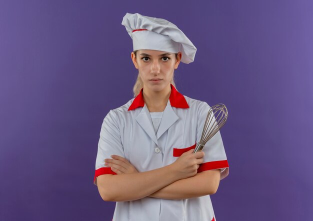 Bastante joven cocinero en uniforme de chef de pie con postura cerrada y sosteniendo batidor mirando