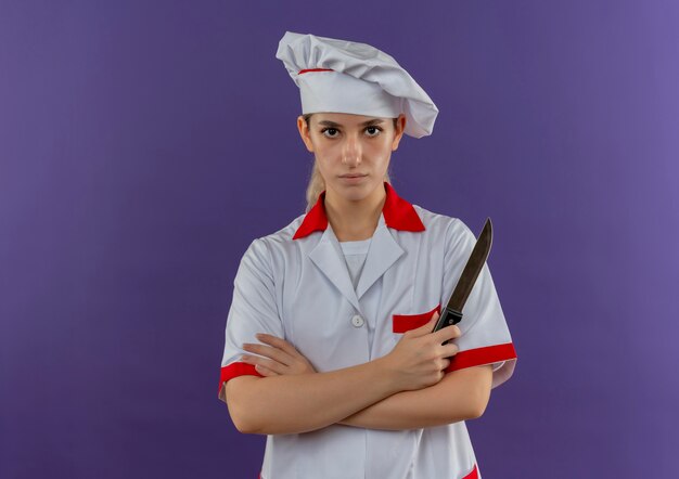 Bastante joven cocinero en uniforme de chef de pie con postura cerrada sosteniendo un cuchillo y mirando