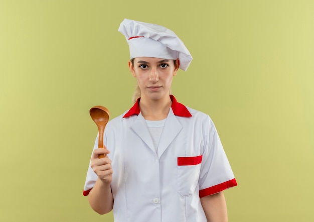 Bastante joven cocinero en uniforme de chef sosteniendo una cuchara y mirando aislado sobre fondo verde