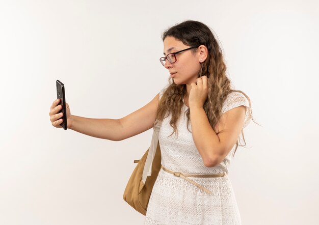 Bastante joven colegiala con gafas y bolsa trasera tomando selfie aislado sobre fondo blanco.