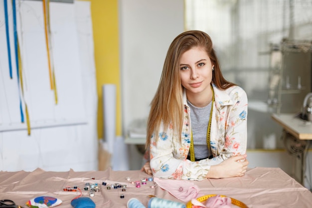 Foto gratuita bastante joven costurera en camisa apoyada en la mesa y mirando soñadoramente a la cámara en un moderno taller de costura