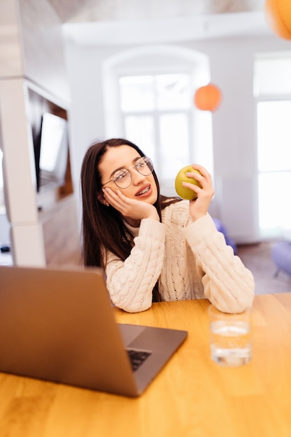 Bastante joven está sentada en la cocina y trabajando en su computadora portátil y teléfono móvil