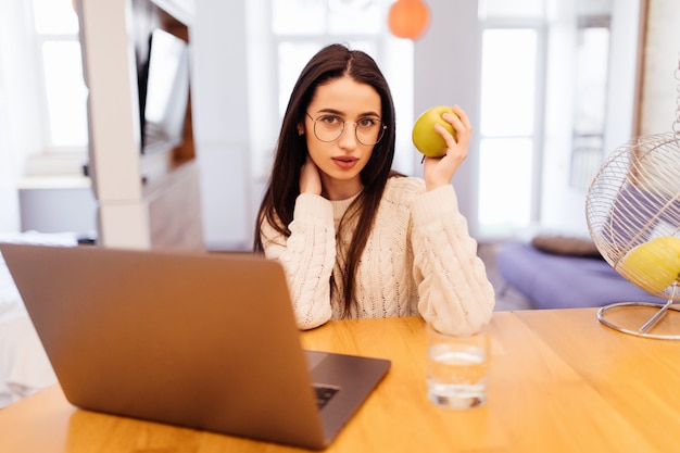 Bastante joven está sentada en la cocina y trabajando en su computadora portátil y teléfono móvil