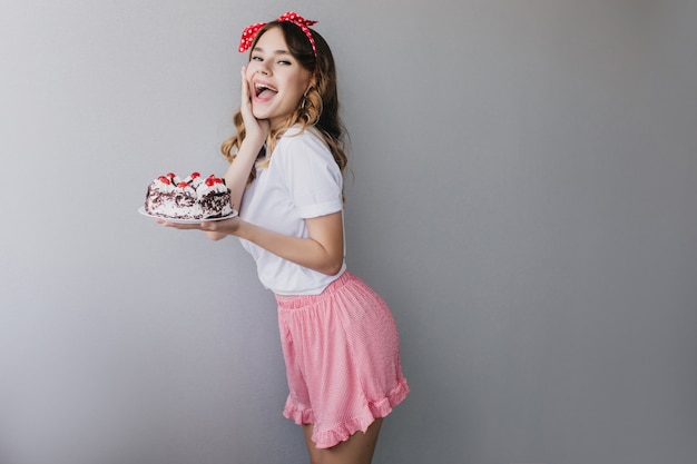 Bastante joven en falda rosa celebrando cumpleaños. Entusiasta chica de cabello oscuro bailando con pastel dulce.