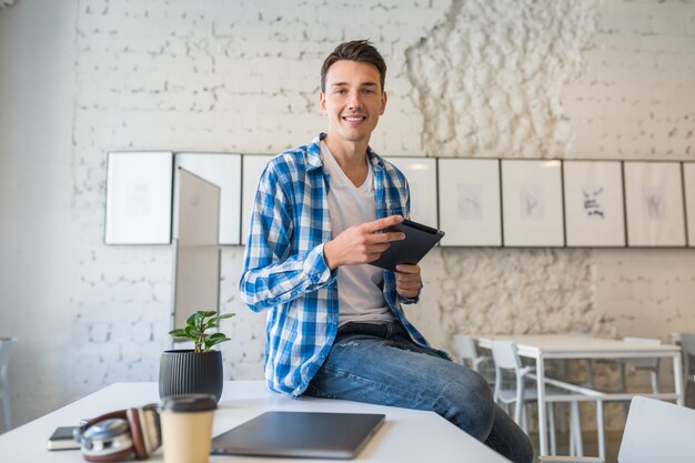 Bastante joven guapo en camisa chekered sentado en la mesa con tablet PC en la oficina de trabajo conjunto,