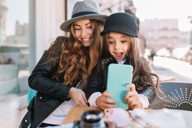 Bastante joven madre y su linda hija se divierten y se toman selfies. niña sorprendida mirando por teléfono y sonriendo en el fondo de la ciudad soleada. familia con estilo, verdadera emoción, buen humor.