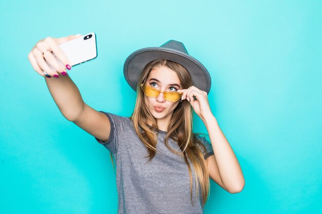 Bastante joven modelo en camiseta de moda, sombrero y gafas transparentes con teléfono en sus manos hace selfie aislado sobre fondo verde