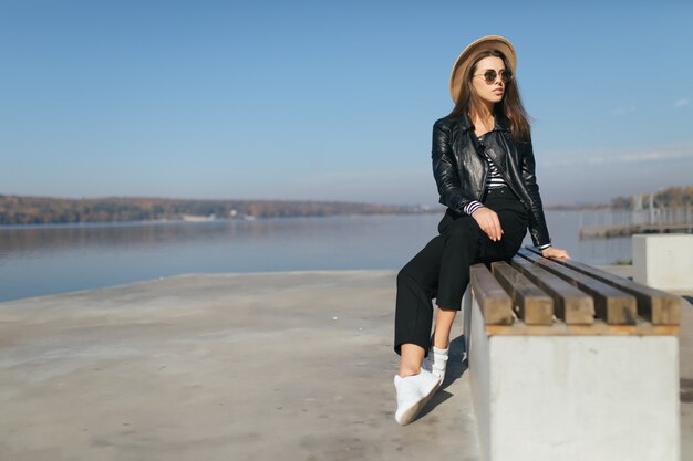 Bastante joven modelo chica mujer posando sentada en el banco en día de otoño en la orilla del lago