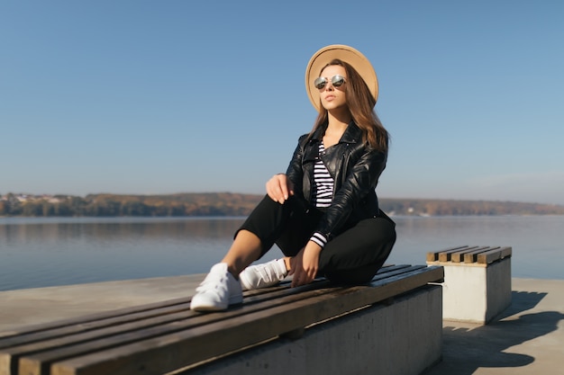 Bastante joven modelo chica mujer sentarse en un banco en día de otoño en la orilla del lago vestida con ropa casual