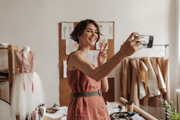 Bastante joven morena de pelo corto en vestido rojo de lino con cinturón negro sonríe, toma selfie, muestra el signo de la paz y posa en la oficina del diseñador de moda