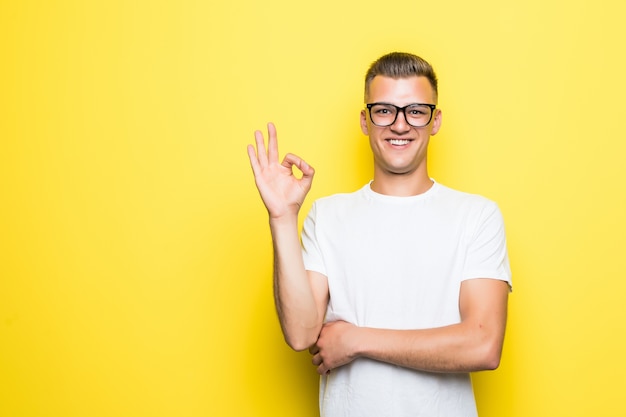 Bastante joven muestra signo bien vestido con camiseta blanca y gafas transparentes aisladas en amarillo