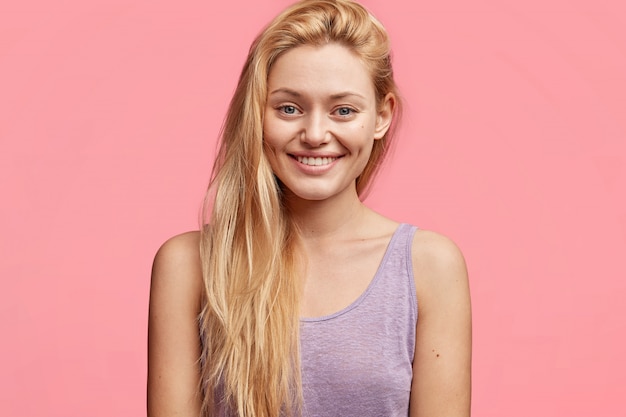 Bastante joven mujer con cabello largo y lujoso y sonrisa agradable, vestida con una camiseta púrpura informal, se regocija con las buenas noticias, posa contra el rosa