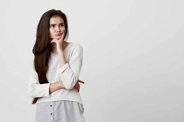 Bastante joven mujer con cabello largo y oscuro mirando a otro lado sosteniendo la mano debajo de la barbilla, construir planes, pensando en algo Pensativa mujer hermosa morena con expresión pensativa y pensativa