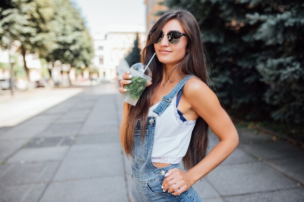 Bastante joven mujer delgada con gafas de sol y camisa en la ciudad tiene una bebida fresca
