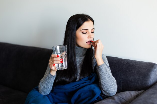 Bastante joven mujer enferma sosteniendo una píldora del día después y un vaso de agua en casa