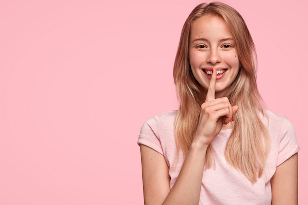 Bastante joven mujer con expresión alegre, le cuenta su secreto a un amigo cercano, hace un signo de silencio, se para contra la pared rosa con espacio de copia a un lado. Concepto de personas, privacidad y secreto.