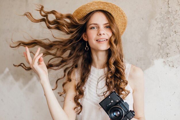 Bastante joven mujer feliz sonriente vistiendo blusa blanca sentada contra la pared con sombrero de paja