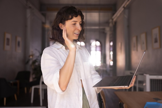 Bastante joven mujer morena caucásica en camisa mira la pantalla de la computadora portátil moderna agitando su mano en el interior