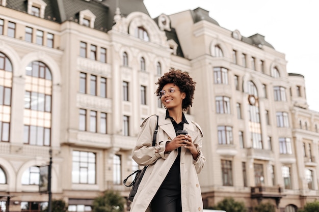 Bastante joven mujer morena feliz en vestido negro y gabardina beige de moda sonríe y sostiene la taza de café al aire libre