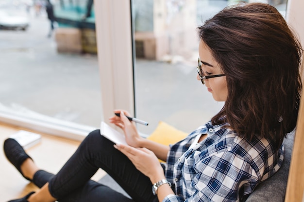 Bastante joven mujer morena con gafas negras sentado en la ventana en casa, escribiendo en el cuaderno. Lugar de trabajo cómodo, análisis, estado de ánimo alegre, estudiante inteligente.