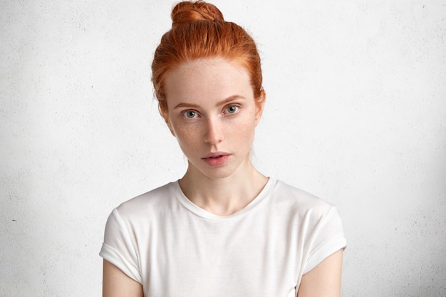 Bastante joven mujer pelirroja en camiseta casual, vestida con camiseta casual, mira con seriedad y confianza a la cámara, tiene un aspecto misterioso