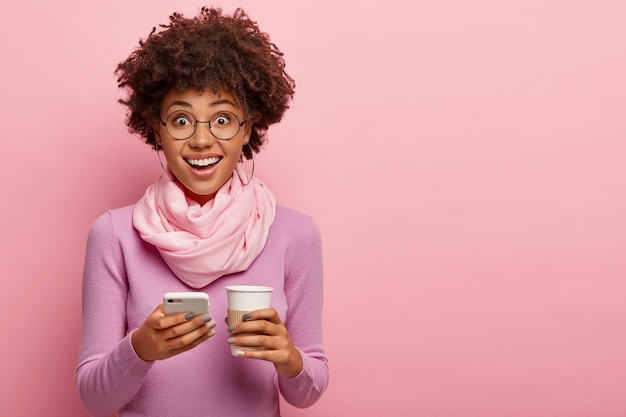 Bastante joven mujer de piel oscura con peinado afro, sostiene un teléfono móvil y una taza de café desechable, vestida con poloneck morado y bufanda, posa sobre una pared rosa. Comunicación, estilo de vida, tecnología