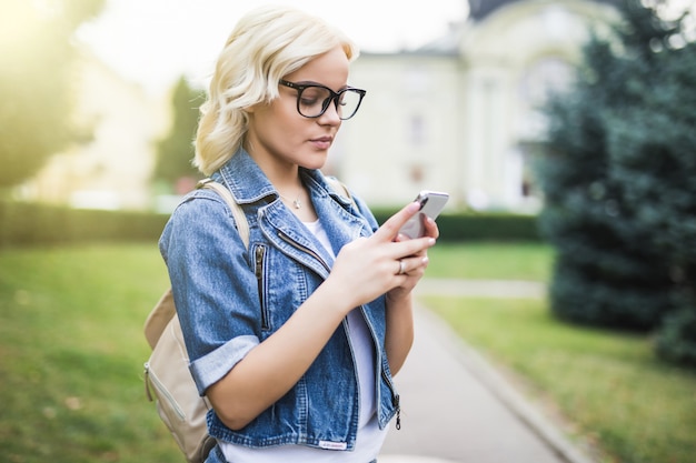 Bastante joven mujer rubia usa el teléfono para desplazarse por la conversación de la red social en la mañana de la plaza de otoño de la ciudad