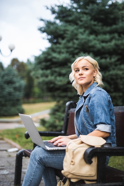 Bastante joven mujer sentada en el banco y usar teléfono y computadora portátil en la ciudad por la mañana de otoño