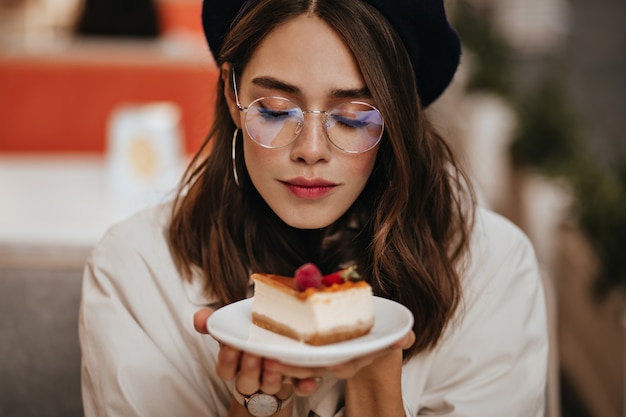 Bastante joven con peinado ondulado oscuro, maquillaje moderno, elegantes pendientes y gabardina beige sentada en la terraza del café de la ciudad y sosteniendo un trozo de tarta de queso