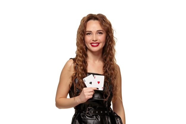 Bastante joven pelirroja o mujer de cabello castaño sonriendo, sosteniendo un par de ases vestidos de látex negro o vestido de cuero aislado sobre fondo blanco. Concepto de casino, industria del juego