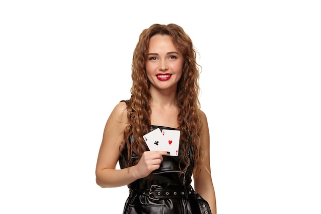 Bastante joven pelirroja o mujer de cabello castaño sonriendo, sosteniendo un par de ases vestidos de látex negro o vestido de cuero aislado sobre fondo blanco. Concepto de casino, industria del juego