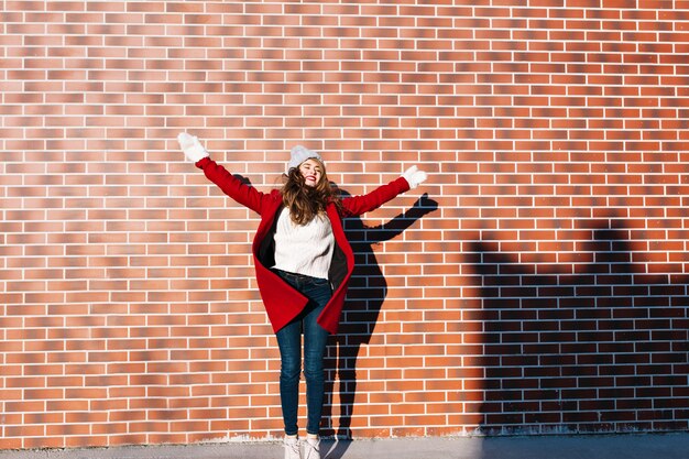 Bastante joven con el pelo largo en abrigo rojo y guantes blancos en la pared exterior. Ella está saltando con los ojos cerrados y una gran sonrisa.