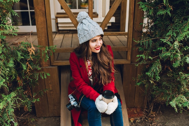 Foto gratuita bastante joven con pelo largo en abrigo rojo sentado en escaleras de madera entre ramas verdes al aire libre. tiene gorro de punto gris, sostiene café con guantes blancos y sonríe a un lado.