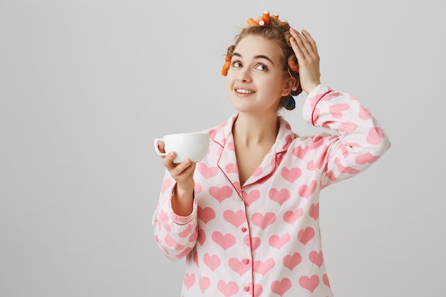 Bastante joven en rizadores de pelo y pijama bebiendo café por la mañana