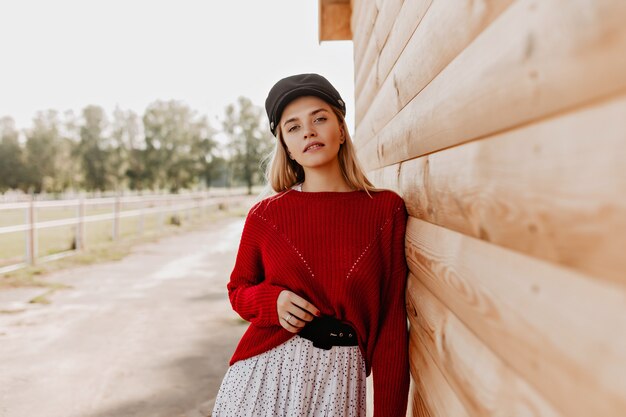 Bastante joven rubia en suéter rojo y sombrero oscuro posando en el parque junto a la pared de madera. Hermosa mujer vestida con ropa de temporada de moda en otoño.