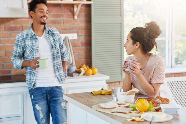 Bastante joven se sienta en la mesa de la cocina, sostiene una taza de té caliente,