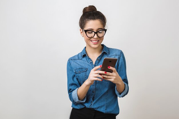 Bastante joven sosteniendo un teléfono inteligente, usando el dispositivo, usando gafas elegantes, sonriendo, aislado, camisa de mezclilla