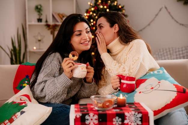 Bastante joven susurra al oído de su amiga sentada en sillones y disfrutando de la Navidad en casa